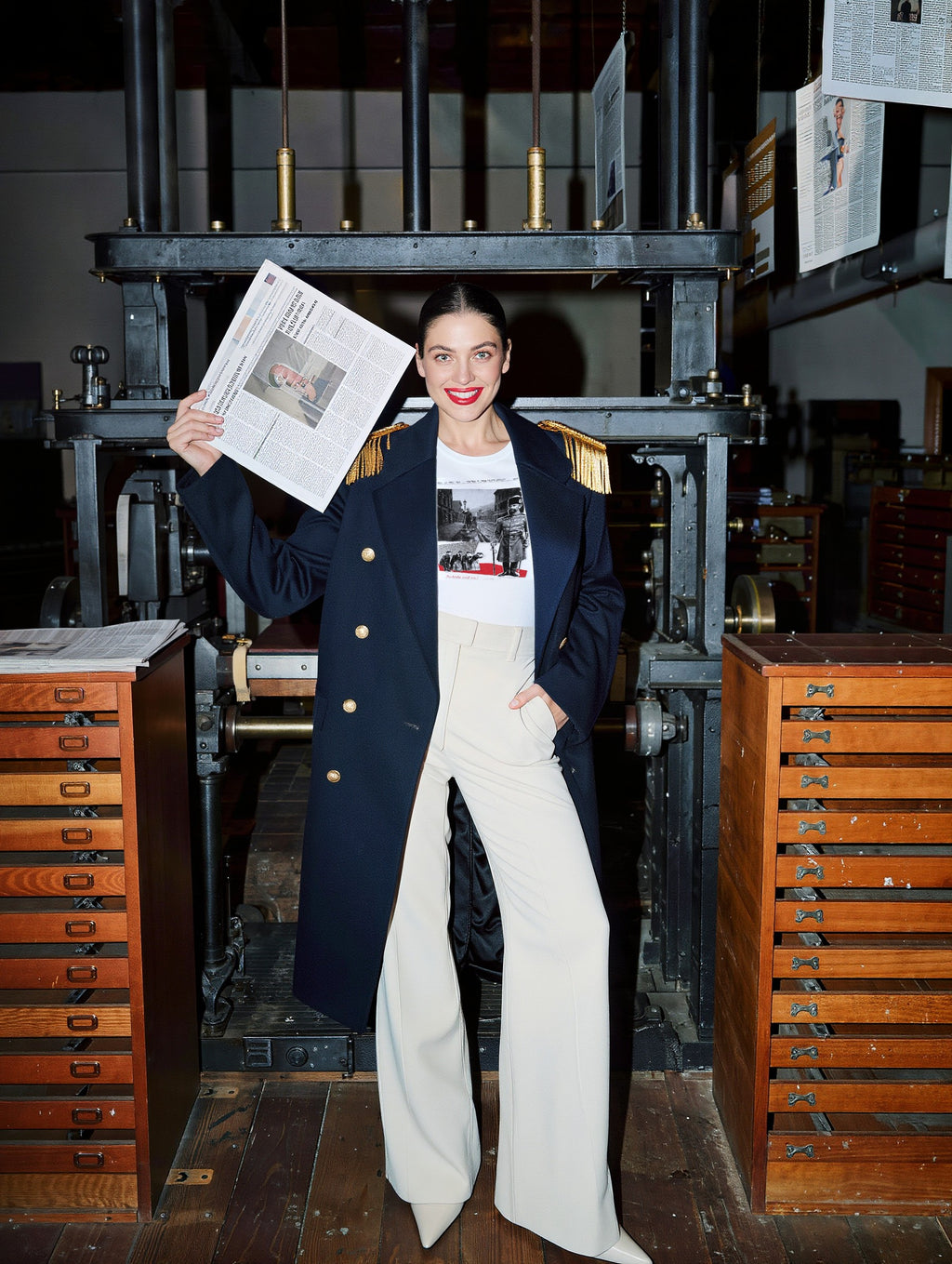Woman in a navy coat in a press room holding a newspaper, wearing Plate XX