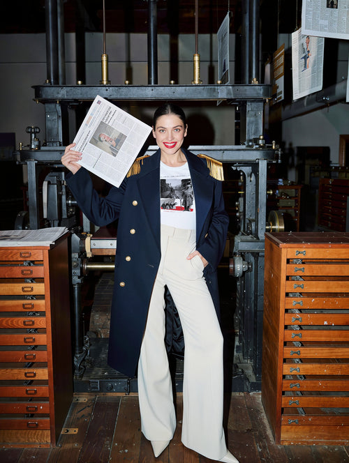 Woman in a navy coat in a press room holding a newspaper, wearing Plate XX